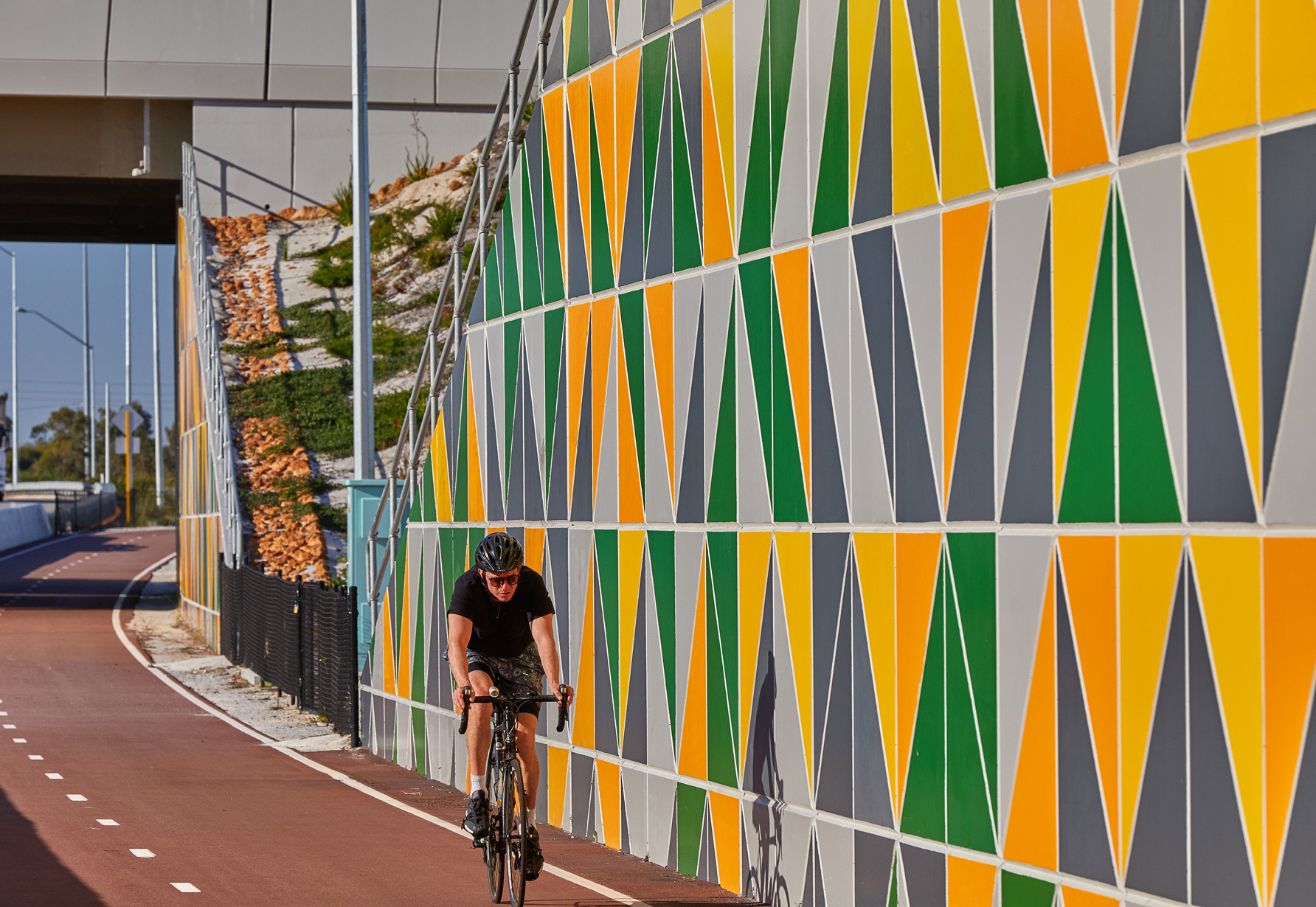 A Reinforced Earth retaining precast concrete wall at Toking Highway, Western Australia, with a colourful modular facing panel. Bicycle rider riding on the cycle path in front of the wall.
